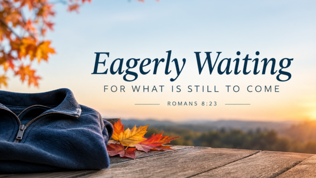 Folded navy blue fleece jacket and autumn maple leaves on a wooden deck at sunset with text reading "Eagerly Waiting FOR WHAT IS STILL TO COME - ROMANS 8:23".